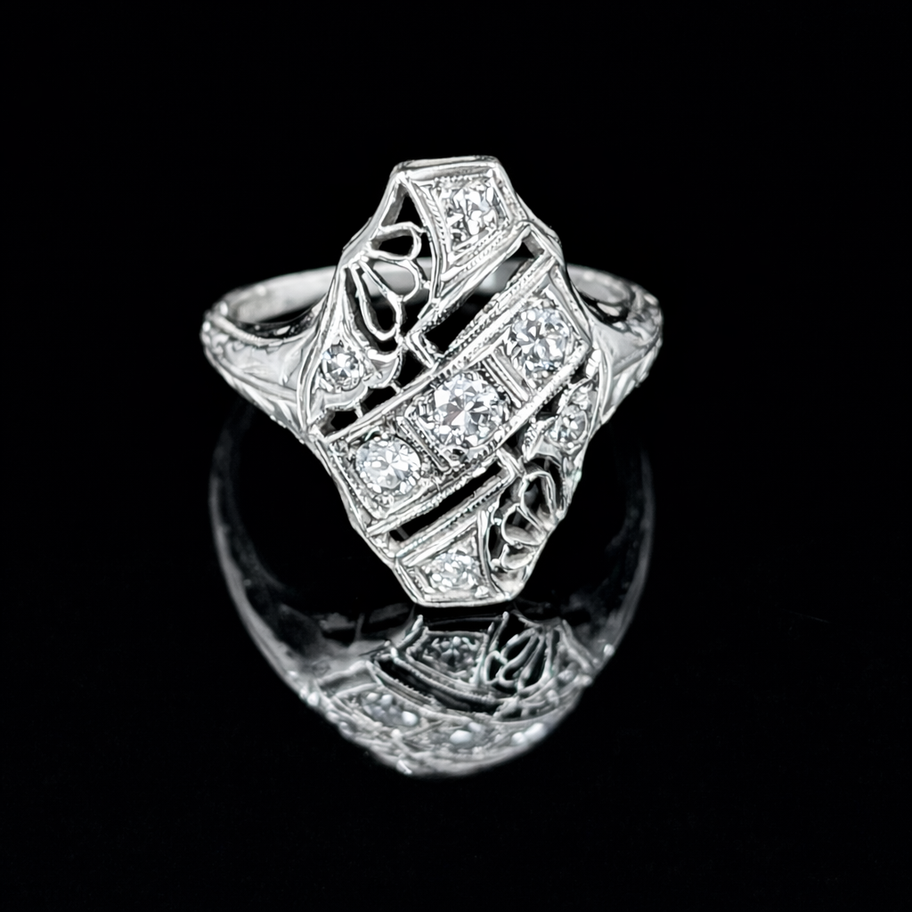 An elegant 18K white gold Art Deco ring with a uniquely shaped, shield-like design. It features intricate geometric openwork filigree and multiple sparkling diamonds set within its detailed structure, presented on a glossy black reflective surface.