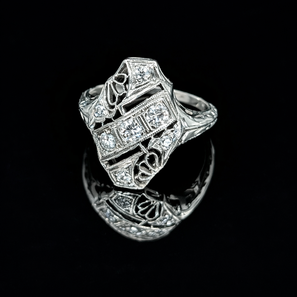 An elegant 18K white gold Art Deco ring with a uniquely shaped, shield-like design. It features intricate geometric openwork filigree and multiple sparkling diamonds set within its detailed structure, presented on a glossy black reflective surface.