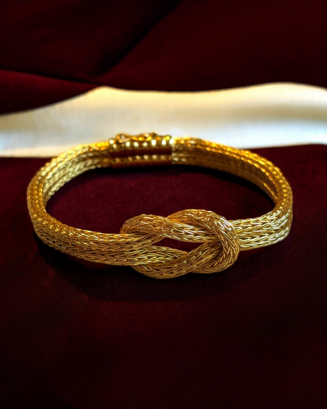 A rich 18K yellow gold Hercules knot bracelet, featuring a prominent woven mesh design and a central intertwined knot. A statement piece with a beautiful design sitting on a deep red velvet jewelry box.