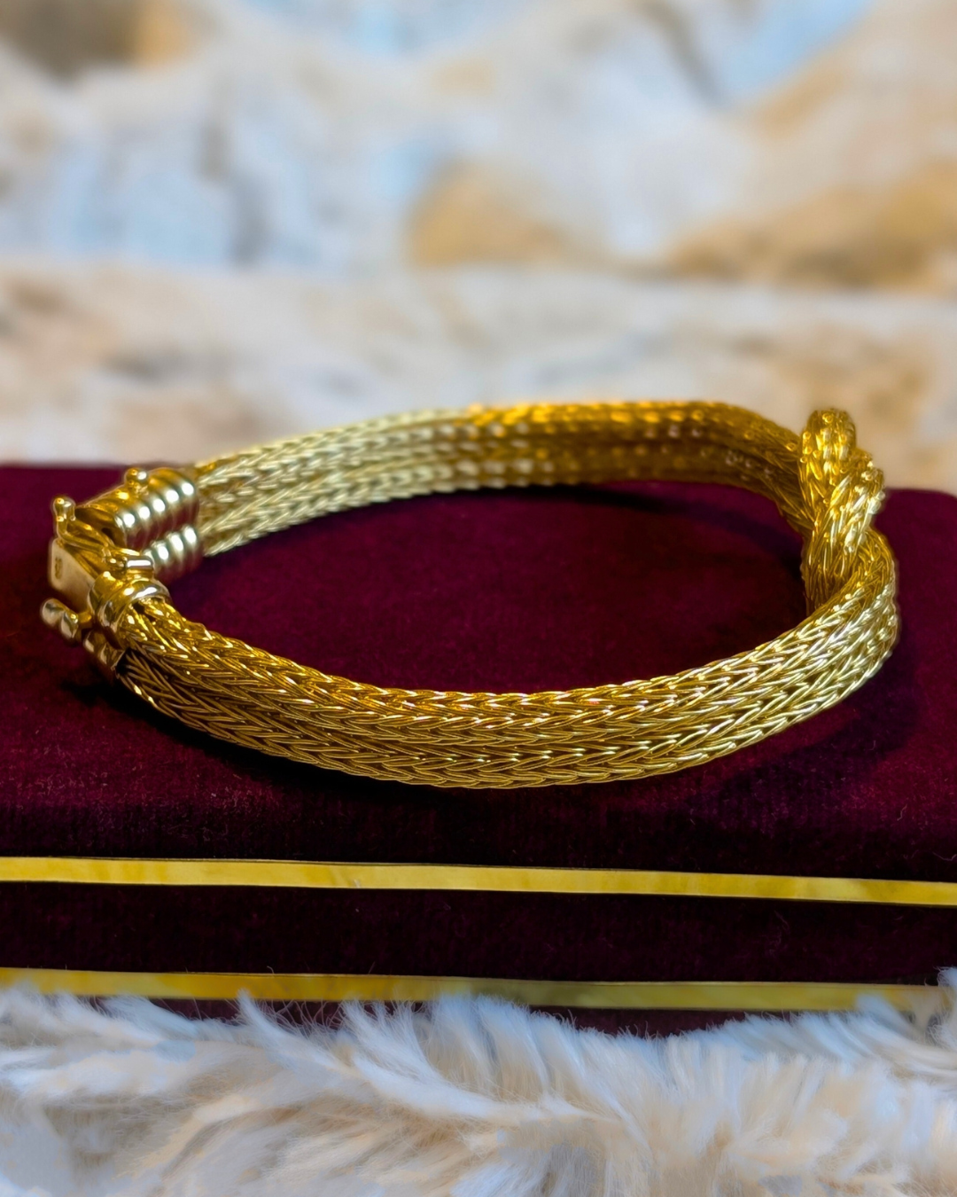 A rich 18K yellow gold Hercules knot bracelet, featuring a prominent woven mesh design and a central intertwined knot. A statement piece with a beautiful design sitting on a deep red velvet jewelry box.