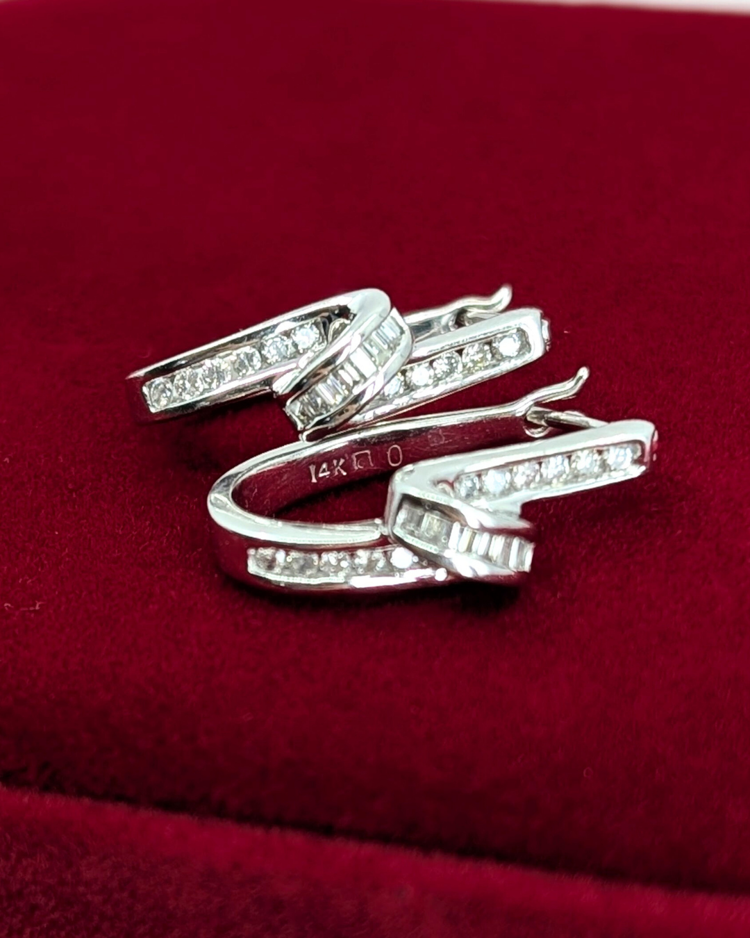 An elegant pair of 14K white gold ribbon earrings on a red velvet surface. Each earring features a curved, polished band with a row of sparkling round and baguette-cut diamonds set along the edge.