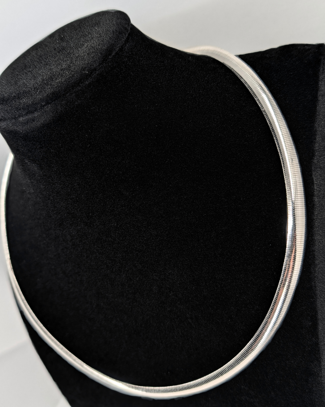 White gold omega collar necklace, showcasing its flat, sleek, and subtly textured surface, displayed on a black neck form.