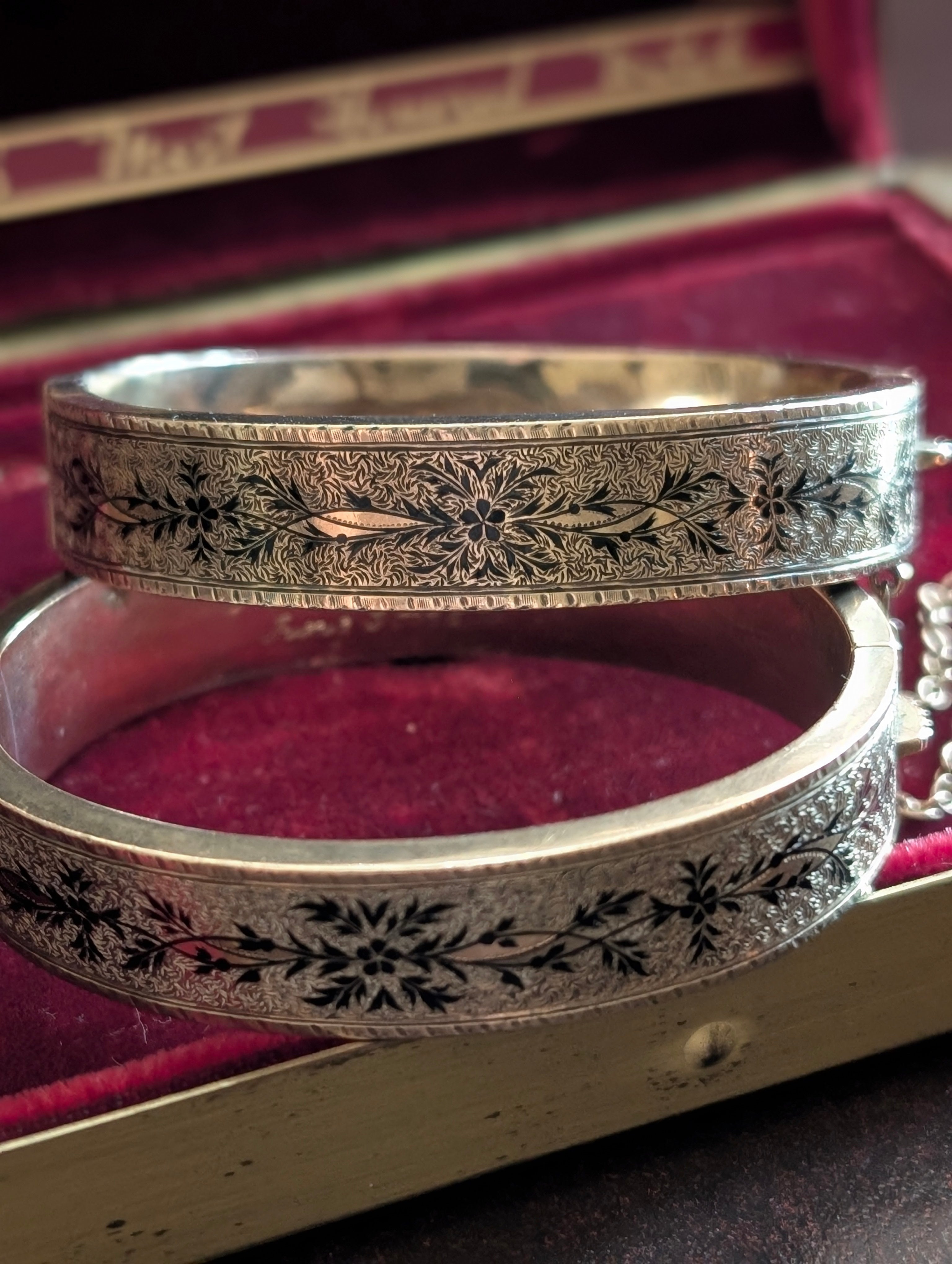 Antique Victorian gold hinged bangles decorated with taille d’épargne black tracery enamel and an 1875 engraved inscription, displayed in a burgundy jewelry box.
